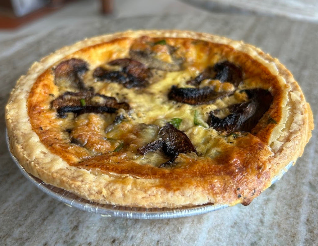 A baked quiche or tart with a golden-brown pastry crust and dark mushroom pieces, served in a small aluminum pie dish on a stone tabletop.